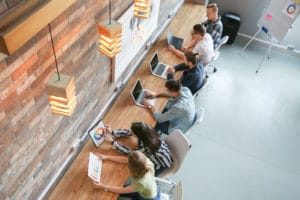 Six people sitting at a long wooden table each working on a laptop or tablet.