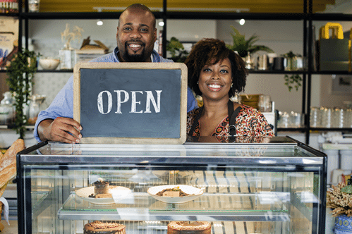 Middle-aged couple standing behind dessert counter holding an open sign. 