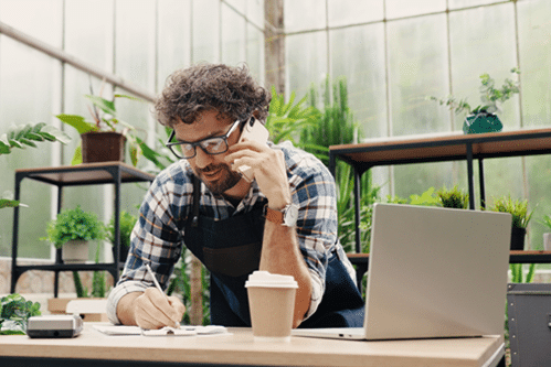 Engaged small business owner on phone with coffee in foreground and greenery in background