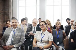 Group of smiling professionals seated in a conference room wearing badges; appears to be attendees at a meeting or seminar.