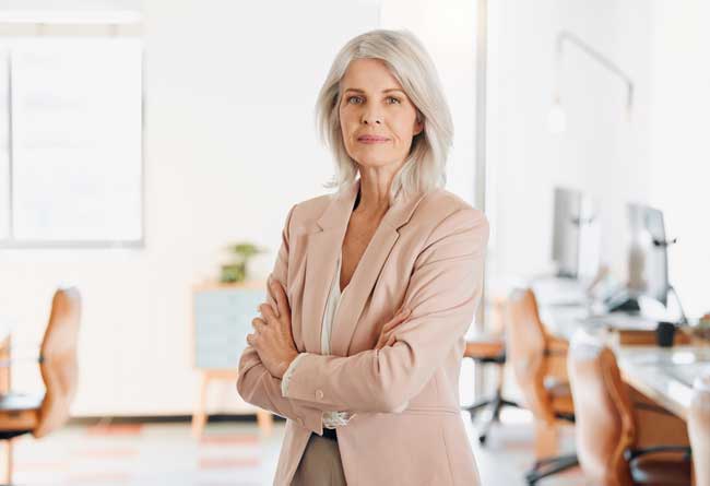 Professional woman with silver hair standing in a bright office, arms crossed, wearing a light pink blazer.