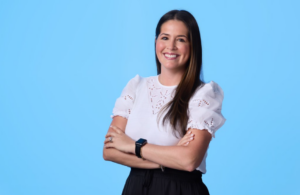 Smiling woman with arms crossed, wearing a white blouse and black skirt, against a blue background.