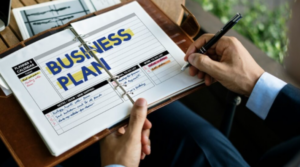 Open binder with a printed 'Business Plan' page and a person writing with a pen on a wooden desk.]","Close-up of a business plan planner being edited, two hands holding a pen over the binder.