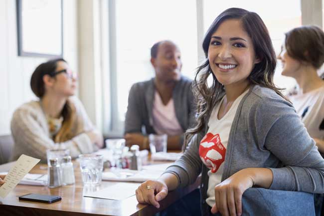 Smiling woman in a gray cardigan sits at a table with colleagues during a meeting in a bright room.